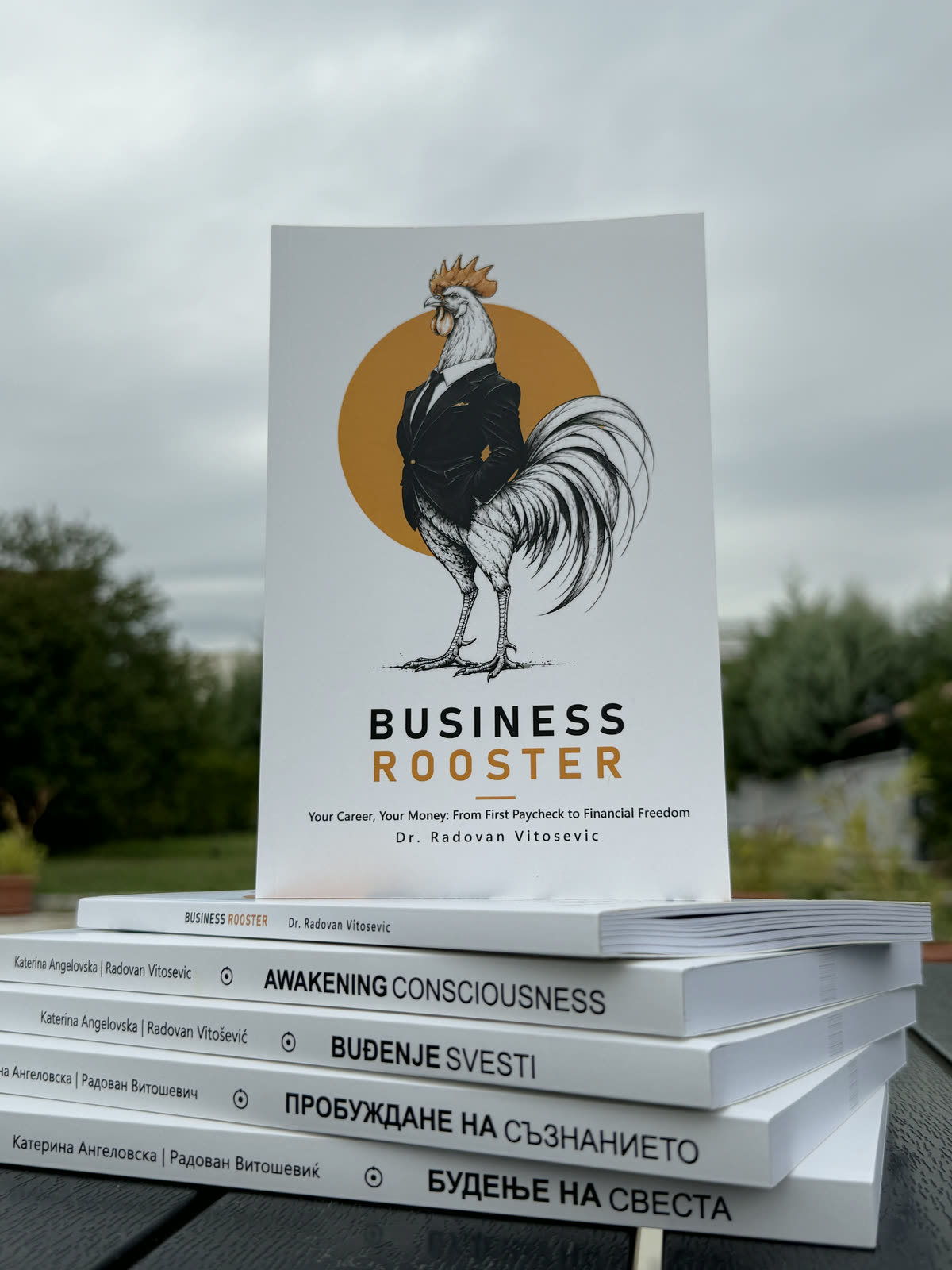 The printed paperback of Business Rooster standing on a stack of Awakening Consciousness in English, Serbian, Bulgarian and Macedonian